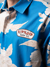 A close-up of a person wearing a blue Surf Co Button Up in size M, featuring large light-coloured leaf patterns and a patch that reads Ripmoid Surf Co. on the chest. Gate21.ee