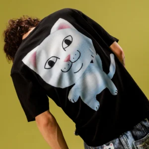 A person with curly hair wears the RIPnDIP NermII Tee in charcoal, which features a large graphic of a cartoon white cat with pink ears, a smiling face, and an upright pose. The person is facing away from the camera. Gate21.ee