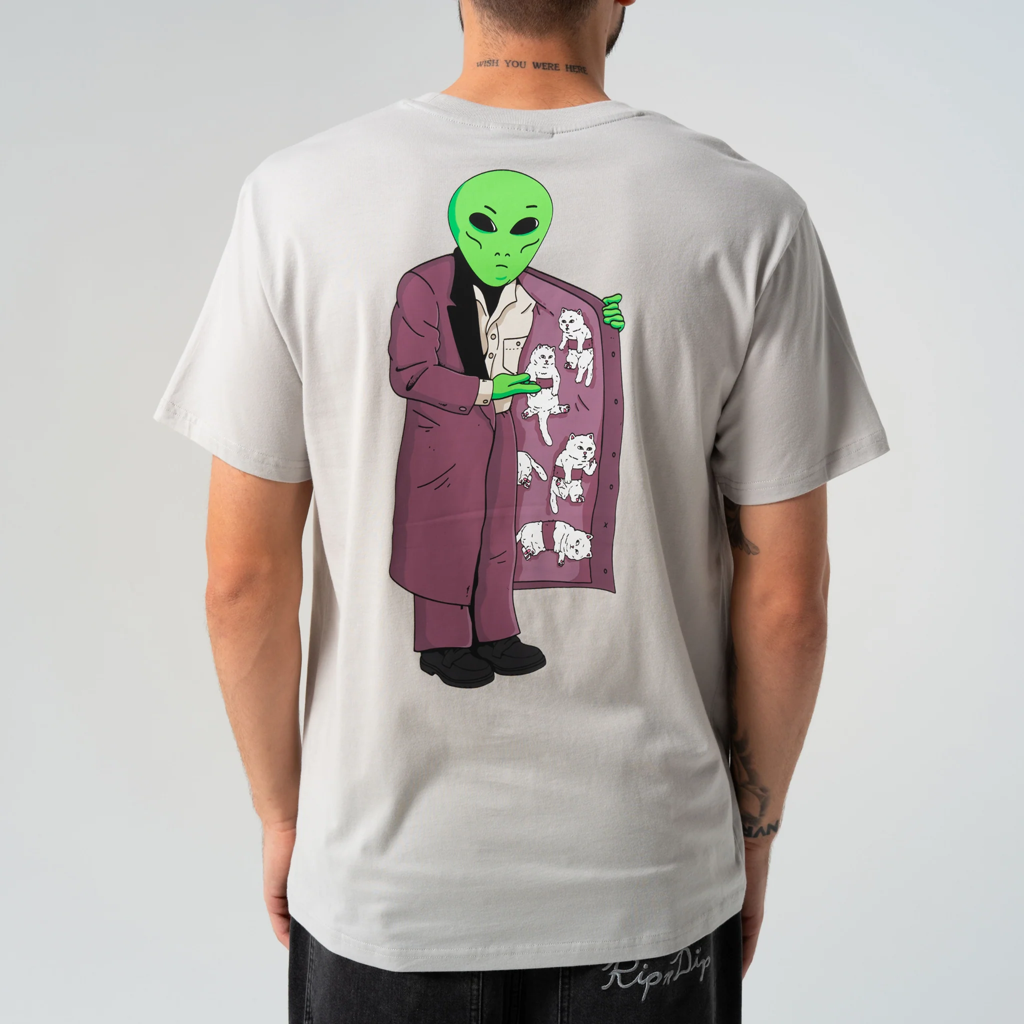 A person wearing the For Sale Pocket Tee - Grey, a light grey t-shirt featuring a playful design in the quirky RipnDip style, with an illustration of an alien in a suit opening a coat to reveal small cartoon cats inside. The individual is shown from the back against a plain background, echoing the fun and imaginative artwork seen on tees like the RIPnDIP Skelly Nerm Smokes Tee Black. Gate21.ee