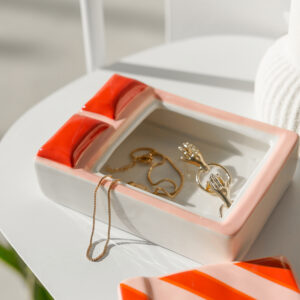 A Bed - Jewellery Box, reminiscent of the dreamy cat jewellery dish by Hello Ferretti, features ceramic with red and pink accents and holds a gold necklace, a ring, and a pair of gold leaf-shaped earrings. The jewellery box rests on a white surface bathed in bright sunlight. Gate21.ee