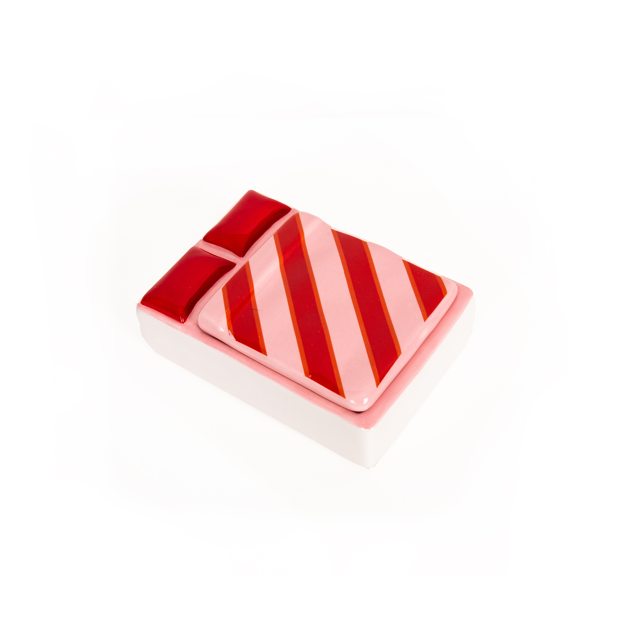 A rectangular ceramic jewellery box called “Bed - Jewellery Box” features a pink base and a lid adorned with red diagonal stripes. At the top, two red rectangular shapes that resemble pillows lend a whimsical touch. The piece is photographed on a white background. Gate21.ee