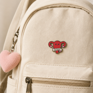 A beige canvas rucksack with a front zip pocket, accessorised with a pink plush heart keyring and the Cuterus - Pin—a red enamel badge shaped like a uterus labelled CUTERUS—attached to the pocket. For extra flair, it also features the iconic This is fine enamel badge, perfect for days when you want your bag to embody both cute and chaotic vibes. Gate21.ee