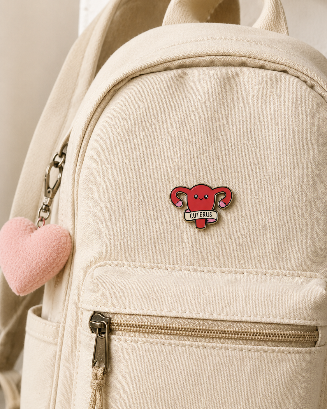 A beige canvas rucksack with a front zip pocket, accessorised with a pink plush heart keyring and the Cuterus - Pin—a red enamel badge shaped like a uterus labelled CUTERUS—attached to the pocket. For extra flair, it also features the iconic This is fine enamel badge, perfect for days when you want your bag to embody both cute and chaotic vibes. Gate21.ee