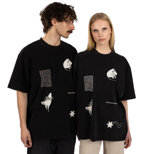 Two people stand side by side wearing the Moomin Heavy Ts - Novels Black, featuring loose black fabric with white Moomin character graphics and text. Both maintain neutral expressions, stylishly showcasing their Moomin Heavy Ts - Novels Black against light backgrounds. Gate21.ee