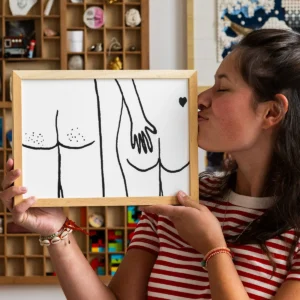 A person in a striped shirt kisses a framed “Poster A4 - Naked Butts,” featuring a black-and-white drawing of two nude figures from behind, with one figure’s hand resting on the other’s buttock. The background displays shelves with various small items, contributing to an intimate atmosphere evocative of Helen B’s distinctive artistic style. Gate21.ee