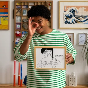 A person in a green and white striped shirt smiles and makes an OK hand gesture while holding the Poster A4 - Bonjour, a framed Helen B drawing of two women—one covering the other’s breasts—featuring the word “Bonjour!” on it. Art and décor are visible in the background. Gate21.ee