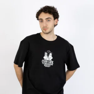 A person with short curly hair wears the Moomin Ts - Club Life Black, a loose black T-shirt featuring two cartoon characters and the text “CLUB LIFE IN MOOMIN VALLEY.” They stand against a plain white background with their hands behind their back. Gate21.ee