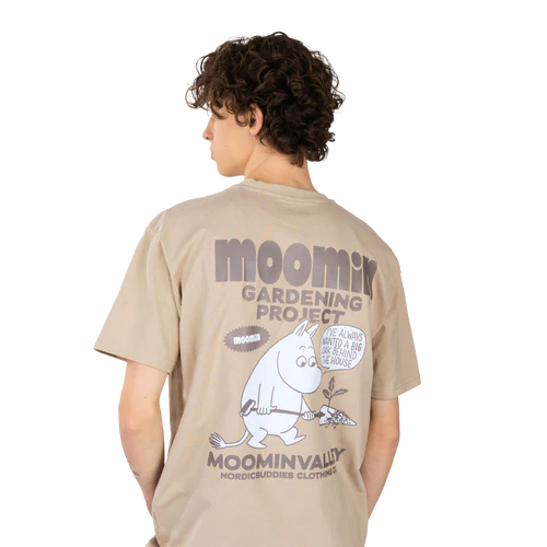 A person with short curly hair wears the charming “Moomintroll Ts – Gardening”—a beige Moomin T-shirt featuring a Moomin character with a spade and plant. The shirt reads: MOOMIN GARDENING PROJECT MOOMINVALLEY NORDICBUDDIES CLOTHING. Gate21.ee