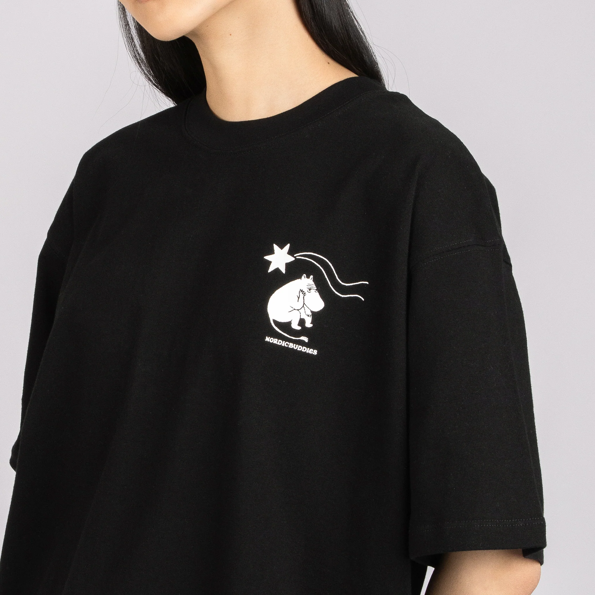 A person with long dark hair, visible from the neck to mid-torso, is wearing the Moomin Heavy Ts - Novels 2 Black. The shirt features a small white graphic of the Moomins and a shooting star on the upper left chest. Gate21.ee