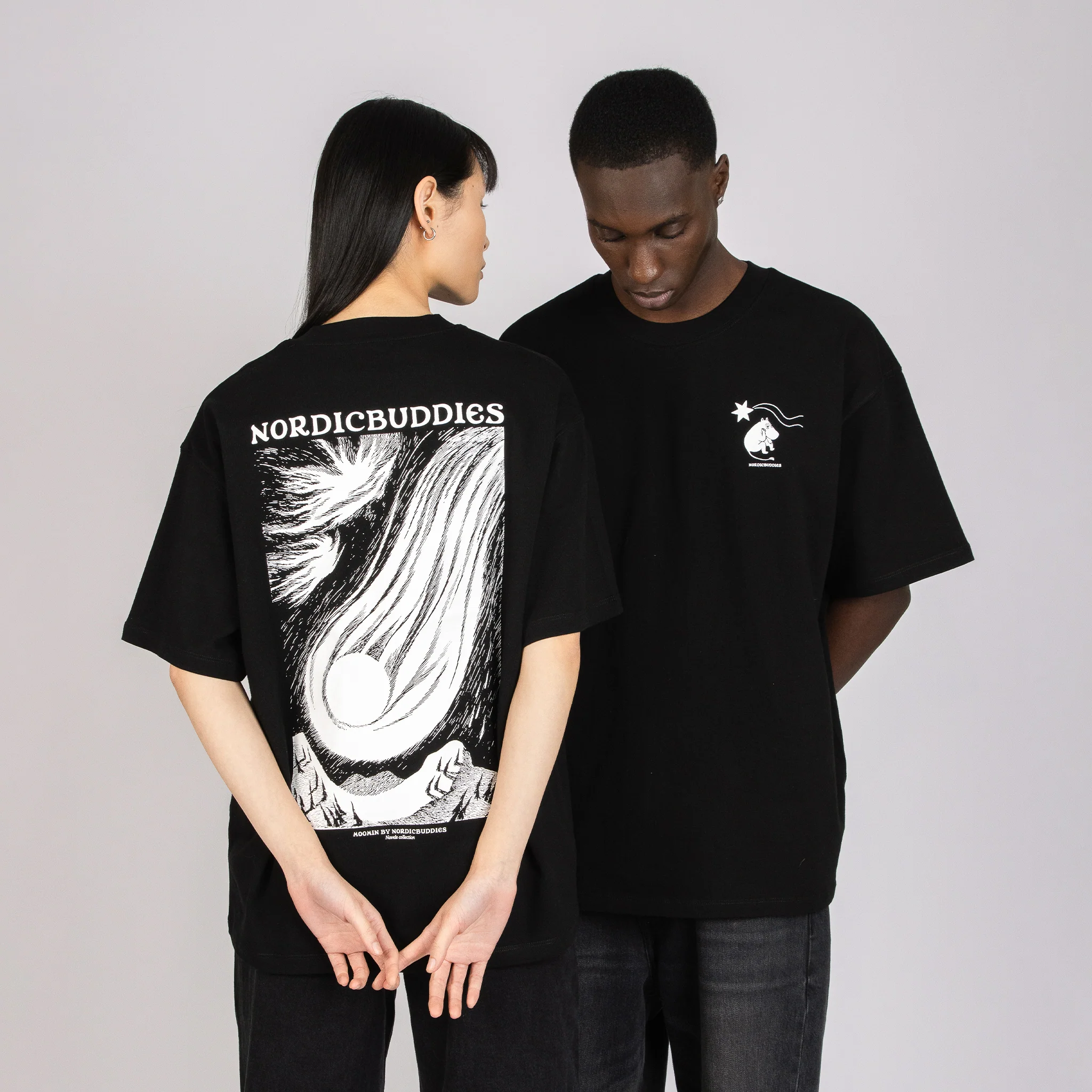 Two people wear the Moomin Heavy Ts - Novels 2 Black T-shirts. One faces forward, showing a small white logo on the chest, while the other faces backwards to display a large illustration and “NORDICBUDDIES” text on the back. Both stand against a plain background. Gate21.ee