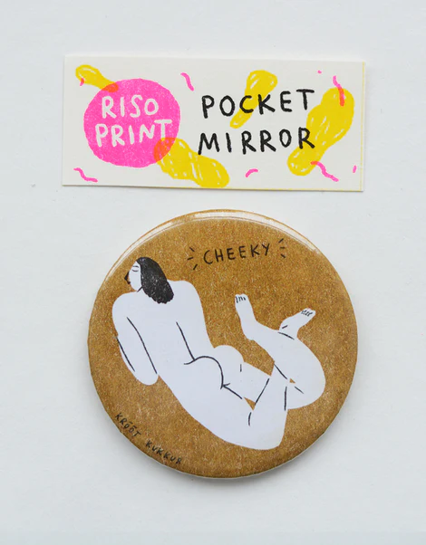 A round pocket mirror, inspired by Krõõt Kukkur Playing Cards – Nudes, features an illustration of a reclining nude figure in Krõõt Kukkur’s playful style with CHEEKY written above. The packaging reads RISO PRINT POCKET MIRROR and is decorated with yellow shapes on a white background, evoking the bold graphics typical of vintage playing cards. Gate21.ee