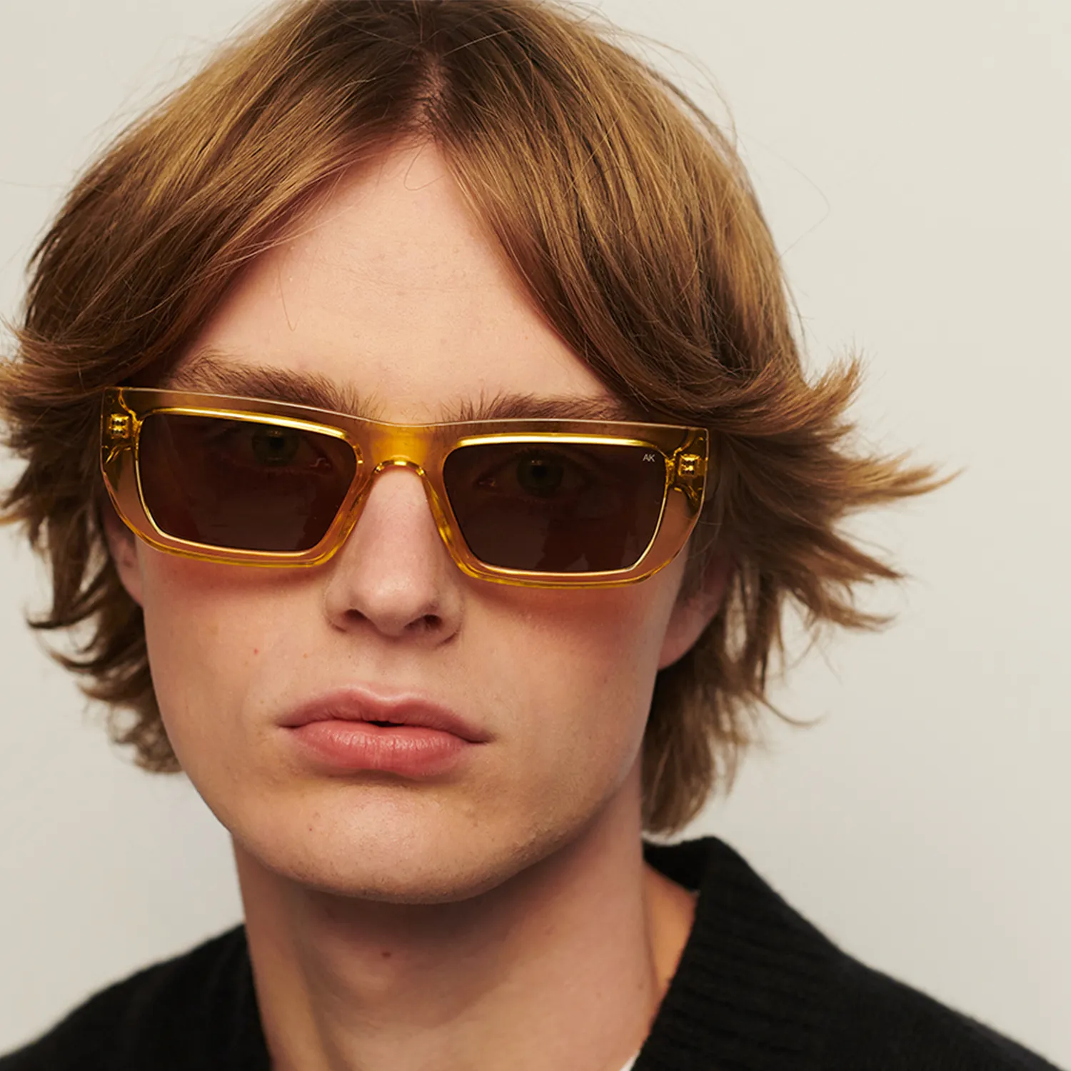 A person with medium-length, light brown hair wears rectangular FAME - Yellow Transparent sunglasses from A. Kjærbede and a black top. The background is plain and neutral. Gate21.ee