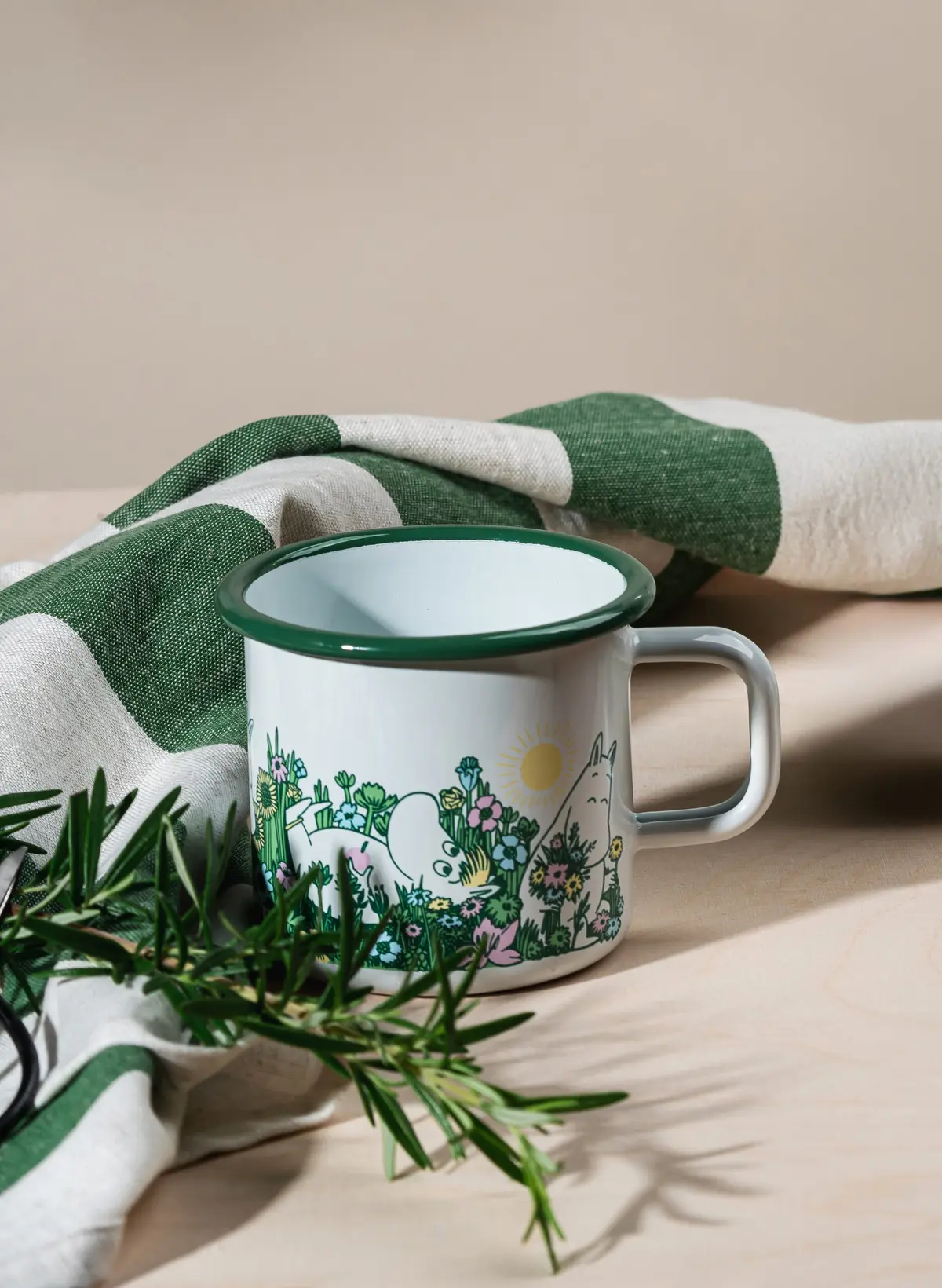 A Garden - Moomin enamel mug 3.7dl, featuring white enamel with green trim and a whimsical cartoon nature scene inspired by classic Moomin designs, sits on a light wood surface beside fresh rosemary sprigs and a green-and-white striped cloth. This charming garden mug offers a generous 3.7dl capacity—perfect for your favourite outdoor sips. Gate21.ee