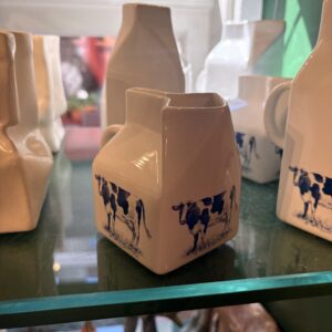 A Creamer LEHM - S, a small ceramic milk jug shaped like a milk carton and decorated with blue cow illustrations on its sides, sits on a glass shelf among other similarly styled ceramic containers. Gate21.ee