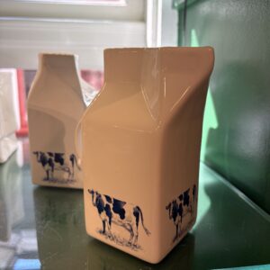 A Creamer LEHM - S, a ceramic jug shaped like a milk carton and adorned with a black and white cow illustration, sits on a glass shelf beside another similar jug as sunlight filters through a nearby window. Gate21.ee