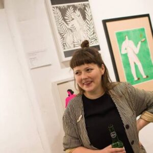 A woman with brown hair in a topknot, wearing a striped jacket and holding a Kr&otilde;&otilde;t Kukkur Enamel Cup - K&Otilde;IK, stands in front of framed artwork on a white gallery wall. She is smiling and looking slightly to the right. Gate21.ee