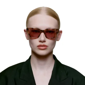 A person with light skin and blonde hair pulled back wears KAWS - Coquina tortoiseshell sunglasses and a black blazer, facing forwards against a plain white background. Gate21.ee