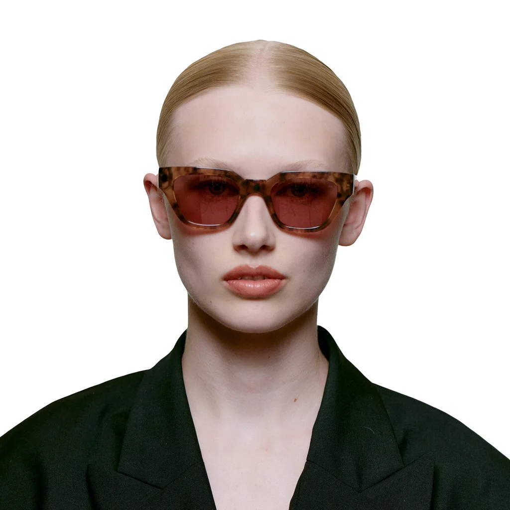 A person with light skin and blonde hair pulled back wears KAWS - Coquina tortoiseshell sunglasses and a black blazer, facing forwards against a plain white background. Gate21.ee