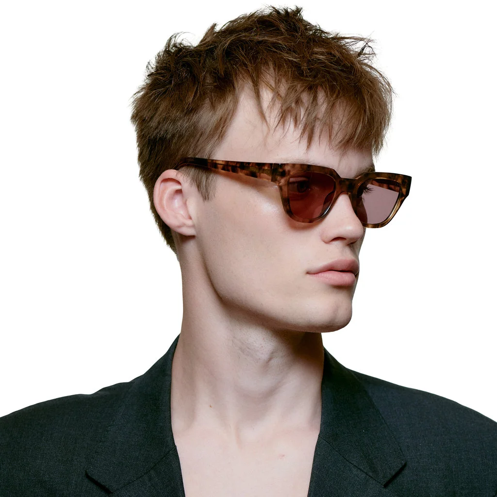 A person with tousled light brown hair wears stylish brown tortoiseshell KAWS - Coquina sunglasses and a dark blazer, looking to the side against a plain white background. Gate21.ee