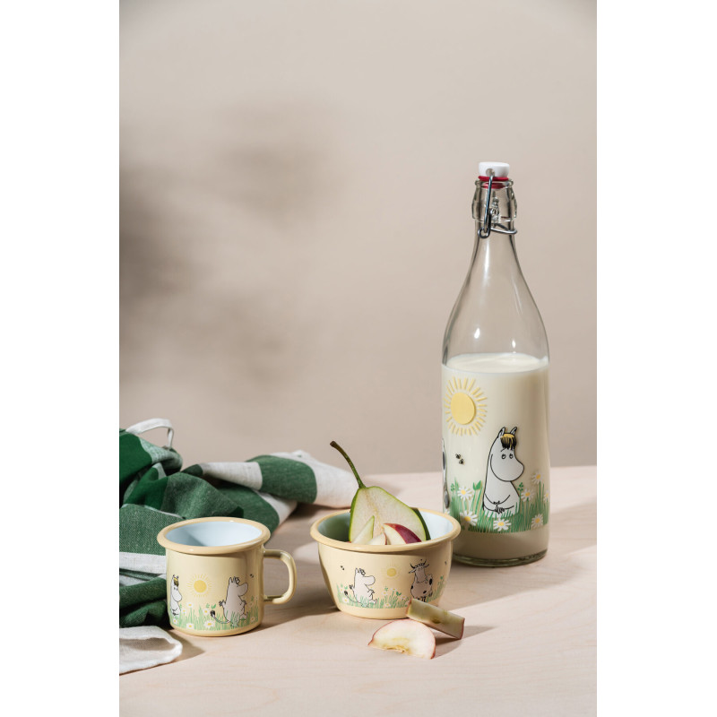 A Meadow – Moomin enamel mug 2.5dl is arranged on a light wooden surface alongside a glass bottle of milk and a bowl with apple slices. Each item features charming cartoon hippo-like Moomin characters and grass motif designs, evoking the classic Moomin enamel collection. A green striped cloth is partially visible in the background. Gate21.ee
