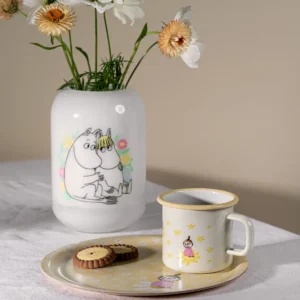 A 3.7dl “Flowers - Moomin” enamel mug sits alongside a ceramic vase decorated with cartoon characters and white flowers, a plate of biscuits, and another cartoon character, all arranged on a white tablecloth against a neutral background. Gate21.ee