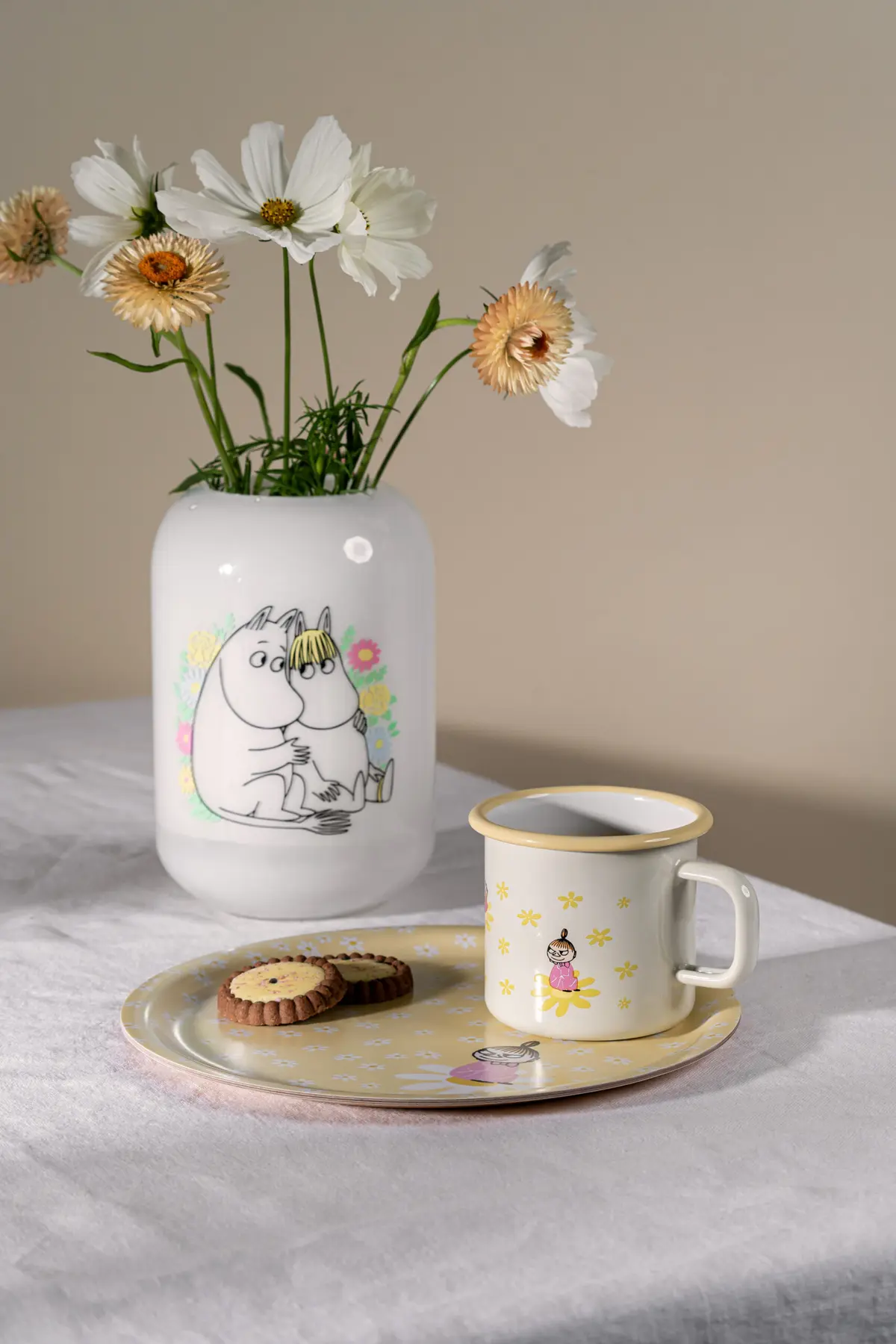 A 3.7dl “Flowers - Moomin” enamel mug sits alongside a ceramic vase decorated with cartoon characters and white flowers, a plate of biscuits, and another cartoon character, all arranged on a white tablecloth against a neutral background. Gate21.ee