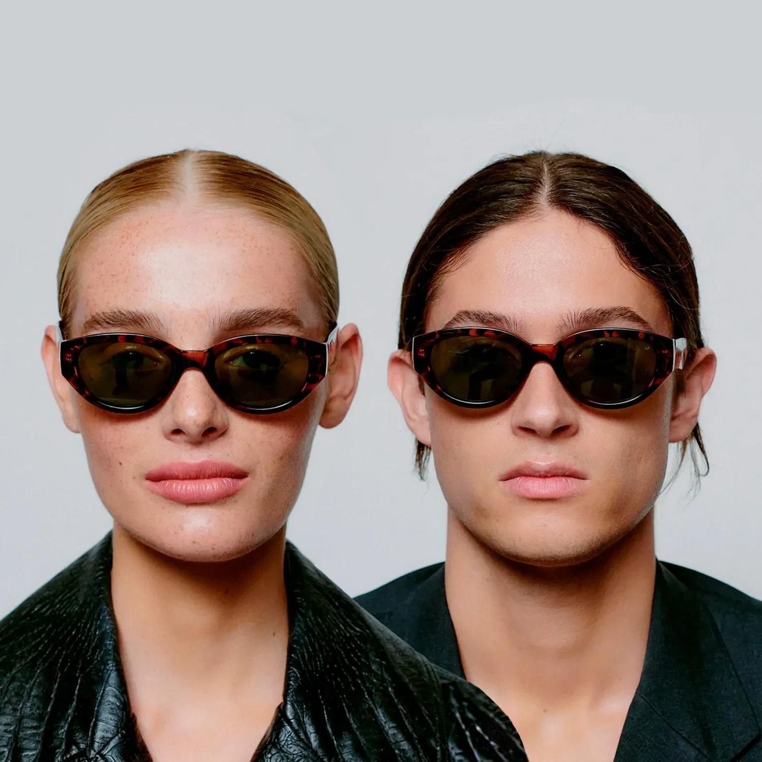 Two people with slicked-back hair and neutral expressions wear Winnie - Demi Tortoise cat-eye sunglasses by A. Kjærbede and dark clothing, standing side by side against a plain light grey background. Gate21.ee