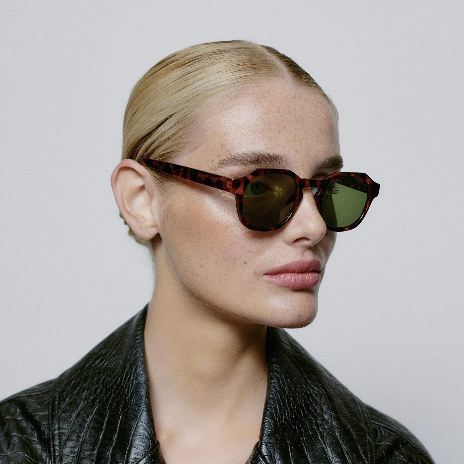 A person with fair hair pulled back wears Zan - Havana sunglasses by A. Kjærbede and a black textured jacket, looking slightly to the side against a plain white background. Gate21.ee