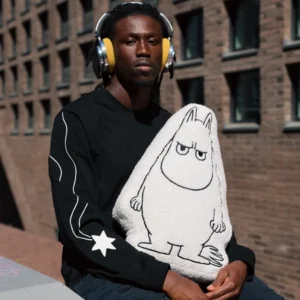 A person wearing yellow headphones and a black sweatshirt with a star on the sleeve sits outdoors, holding a Moomintroll Temper Cushion&mdash;a plush toy featuring the white, pointy-eared character&mdash;with a brick building in the background. Gate21.ee