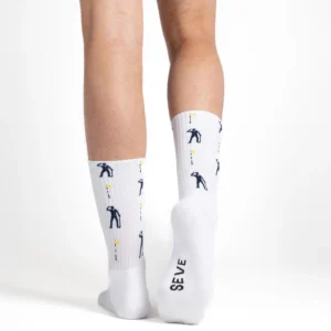 A person’s lower legs wearing The Game - Athletic white Jimmy Lion socks, which feature golfer graphics and yellow flags. The sole of the right sock displays the text SEVE. The background is plain white. Gate21.ee