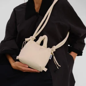 A person wearing a black coat holds an ÖLEND Micro Ona Soft Bag in Butter Yellow—a small, fabric handbag with knotted handles and a shoulder strap. The photo is cropped above the nose, obscuring the person’s full face. Gate21.ee