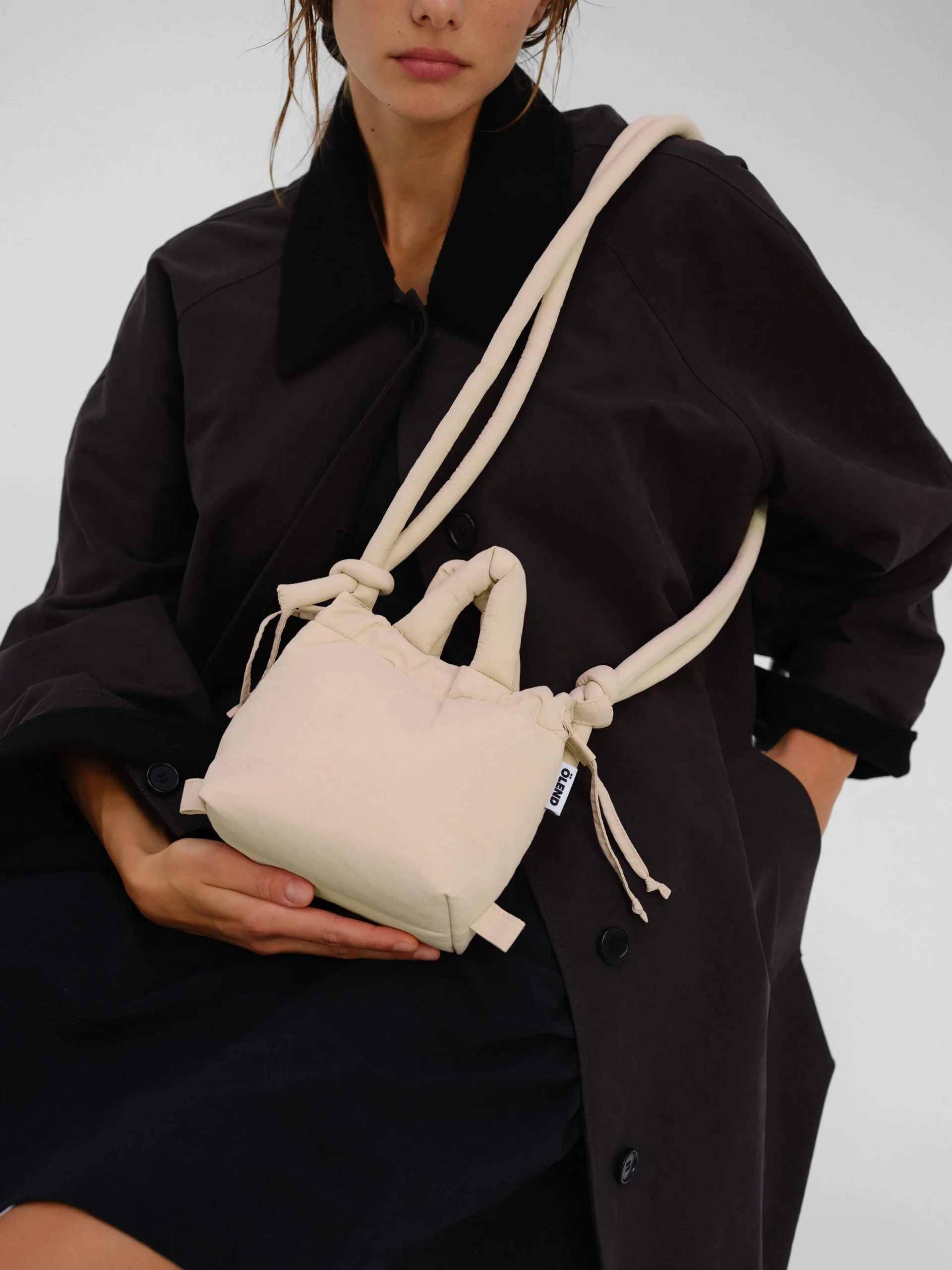 A person wearing a black coat holds an ÖLEND Micro Ona Soft Bag in Butter Yellow—a small, fabric handbag with knotted handles and a shoulder strap. The photo is cropped above the nose, obscuring the person’s full face. Gate21.ee