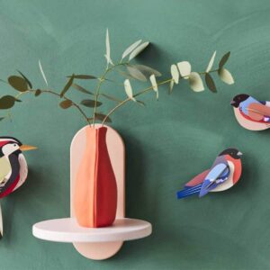 A red vase with a white base holds green branches on a wall. Next to it are three colourful “Finches” by StudioROOF, all mounted on a green background, bringing a playful touch reminiscent of the parasaul family. Gate21.ee