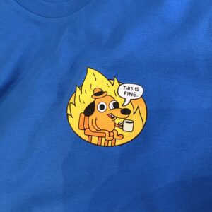 A blue Pinz - T-sargid - K&Otilde;IK shirt showcases a cartoon dog calmly sitting in a chair amid flames, holding a cup with the phrase &ldquo;This is fine&rdquo;&mdash;an ideal choice for fans of the iconic This is fine enamel badge. Gate21.ee