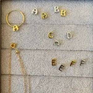 Gold and silver letter charms featuring A, B, C, D, and E are elegantly arranged on a grey jewellery tray alongside ZEN 925 Sterling Silver - Alphabet Earrings. A ring and a necklace chain are also displayed in the top left and bottom left corners for an added touch of ZEN. Gate21.ee