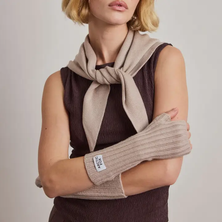 A woman stands with arms crossed, wearing a sleeveless dark dress, a beige jumper draped over her shoulders, and matching Merino Wool Fingerless Gloves in Soft Taupe with a visible Arctic Fox label. The background is plain. Gate21.ee