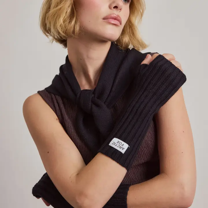 A person with short blonde hair wears a brown sleeveless top, a black knitted scarf draped over their shoulders, and black Merino Wool fingerless gloves, featuring a distinctive white fabric tag. Gate21.ee