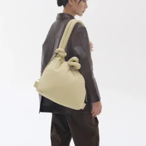 A person wearing a dark brown leather jacket and trousers stands sideways, carrying the ÖLEND Ona Soft Bag in Butter Yellow with knotted handles over their shoulder against a plain white background. Gate21.ee