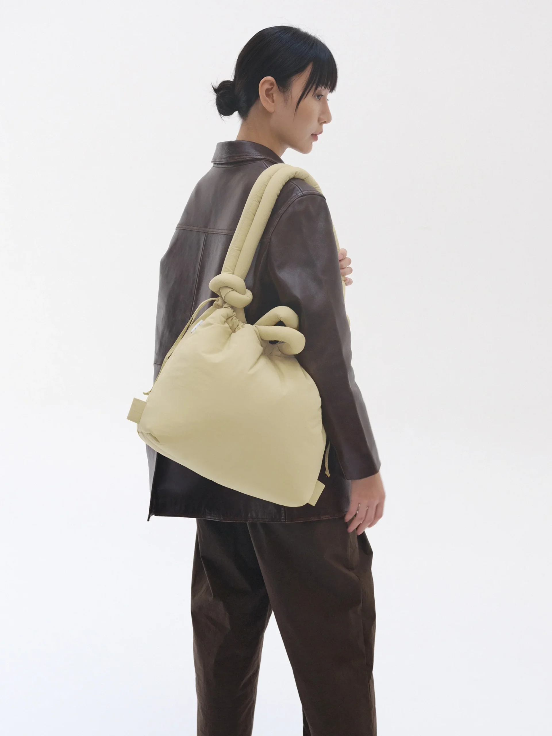 A person wearing a dark brown leather jacket and trousers stands sideways, carrying the ÖLEND Ona Soft Bag in Butter Yellow with knotted handles over their shoulder against a plain white background. Gate21.ee
