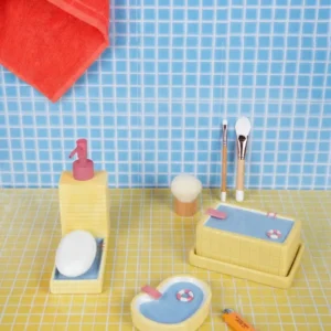 A Pool - Butter Dish from L’expressionist gifts, shaped like a mini bath, is arranged on a yellow tiled surface. A red towel hangs on a blue tiled wall in the background, with an orange label lying nearby. Gate21.ee