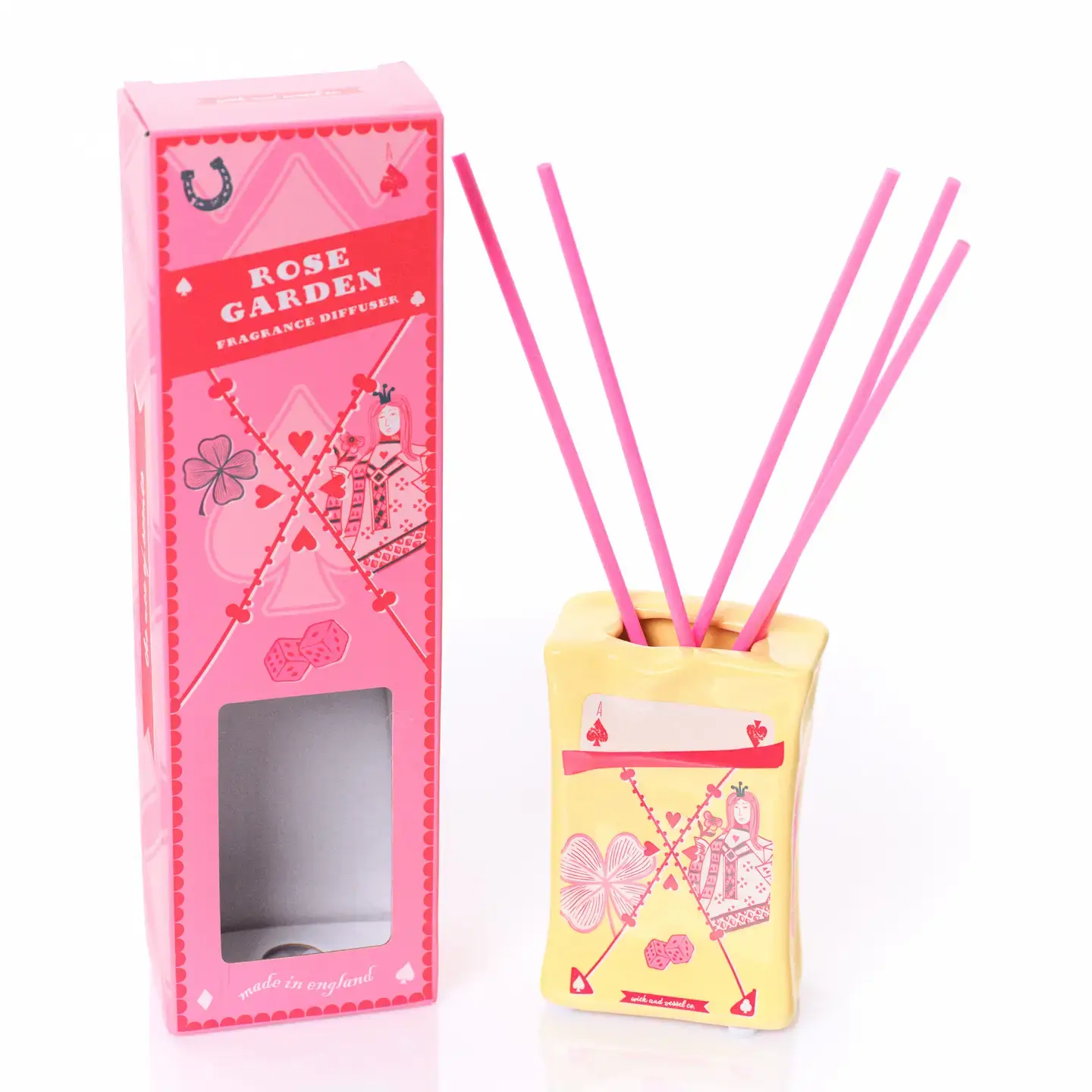 A yellow ceramic Happy Cat - 100ml Diffuser with pink sticks stands next to a pink box labelled Rose Garden. Both are adorned with playful designs, including playing cards and clover symbols, capturing the whimsical charm of Wick & Vessel home fragrance. Gate21.ee