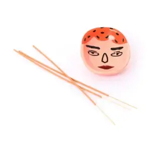 Three incense sticks rest beside the Frank Faces - Incense Burner, a small ceramic holder shaped like a round face with a red and black patterned top and a neutral expression, creating an inviting ambience that echoes the thoughtful design characteristic of Wick & Vessel home fragrance. Gate21.ee
