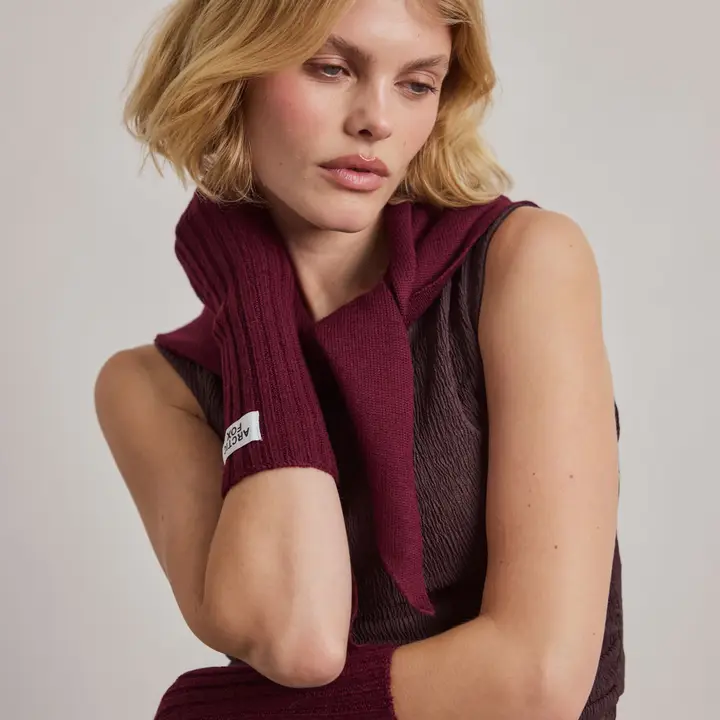 A woman with chin-length blonde hair wears a sleeveless textured top and a burgundy jumper draped over her shoulders, complemented by matching Merino Wool Fingerless Gloves in Cherry Plum. She gazes downward, her hand resting at her neck. Gate21.ee