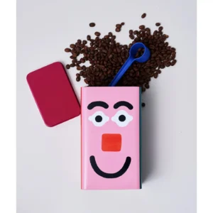 A pink notebook with a cartoon face, a red rectangular object, Kitchen Storage / Coffee Tin - Faces tins, coffee beans spilling out, and a blue spoon are arranged on a white surface. The coffee beans appear to be the hair of the notebook face. Gate21.ee