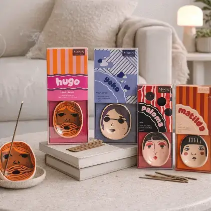 Five boxes of Yoko Faces - Incense Burner products from Wick & Vessel home fragrance, each featuring illustrated faces and names on the packaging, are arranged on a table. One incense coil is burning in a holder, with several sticks placed nearby. A sofa and decor can be seen in the background. Gate21.ee