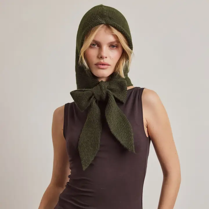 A person wearing a dark sleeveless top and The Bonnet - Forest Green - Recycled stands against a plain light background, facing the camera with a neutral expression. Their hands are tucked snugly into Arctic Fox Reykjavik hand warmers, adding both comfort and style to the ensemble. Gate21.ee