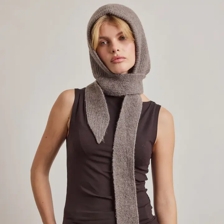 A person wearing a sleeveless black top and The Bonnet in Earthly Grey (Recycled) stands against a plain light background, looking at the camera with a neutral expression, their hands tucked into Arctic Fox Reykjavik hand warmers for added warmth. Gate21.ee