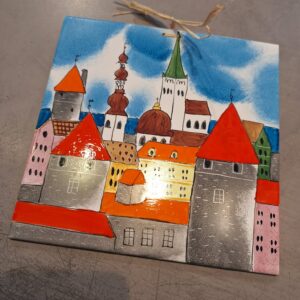 The Helina Tilk Suur alus is a vibrant tile featuring a hand-painted cityscape with striking red-roofed towers, assorted church spires, and a vivid blue sky. Designed with a string for easy hanging, this piece rests elegantly on a grey surface. Gate21.ee