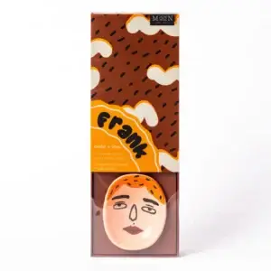 The Frank Faces - Incense Burner includes cedar and lime incense sticks accompanied by a handmade ceramic holder adorned with a simple face illustration. Housed in packaging featuring abstract brown and orange designs, this Wick & Vessel set serves as a chic home fragrance accessory for any room. Gate21.ee