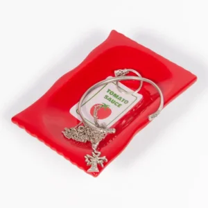 A red Ketchup - Plate with wavy edges displays a silver bracelet, a necklace featuring a figure pendant, and a keyring labelled TOMATO SAUCE with a tomato illustration—distinctive L’expressionist gifts—set against a white background. Gate21.ee